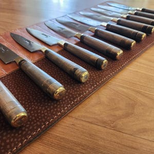 May include: A brown leather knife roll with 12 knives with wooden handles and gold accents. The knives are arranged in a row with the blades facing up.