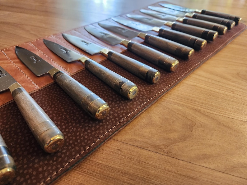 May include: A brown leather knife roll with 12 knives with wooden handles and gold accents. The knives are arranged in a row with the blades facing up.