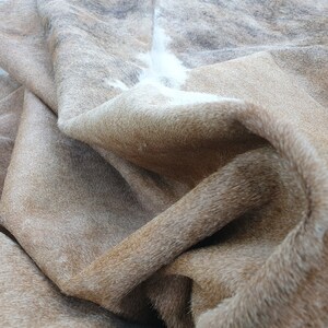 May include: A brown and white cowhide rug with a soft, shaggy texture. The rug is folded and draped, showcasing the natural markings and variations in the hide.