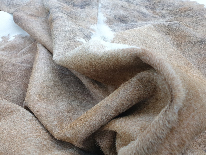 May include: A brown and white cowhide rug with a soft, shaggy texture. The rug is folded and draped, showcasing the natural markings and variations in the hide.