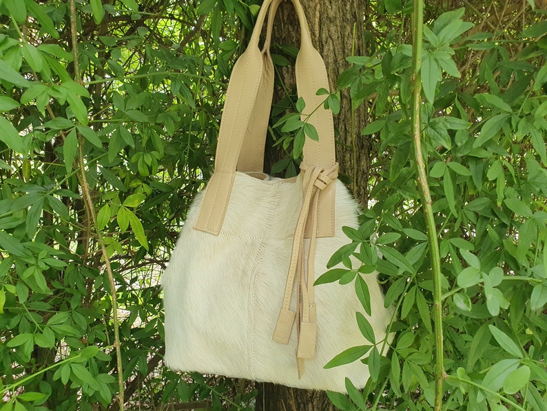 May include: A white leather tote bag with tan leather straps and tassels. The bag is hanging from a tree branch.