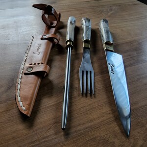 Puede incluir: Un juego de tres utensilios de cocina plateados con mangos de madera. El juego incluye un cuchillo, un tenedor y una brocheta. Los utensilios est&aacute;n dispuestos sobre una superficie de madera. El cuchillo y el tenedor tienen la palabra "Uruguay" grabada en la hoja.