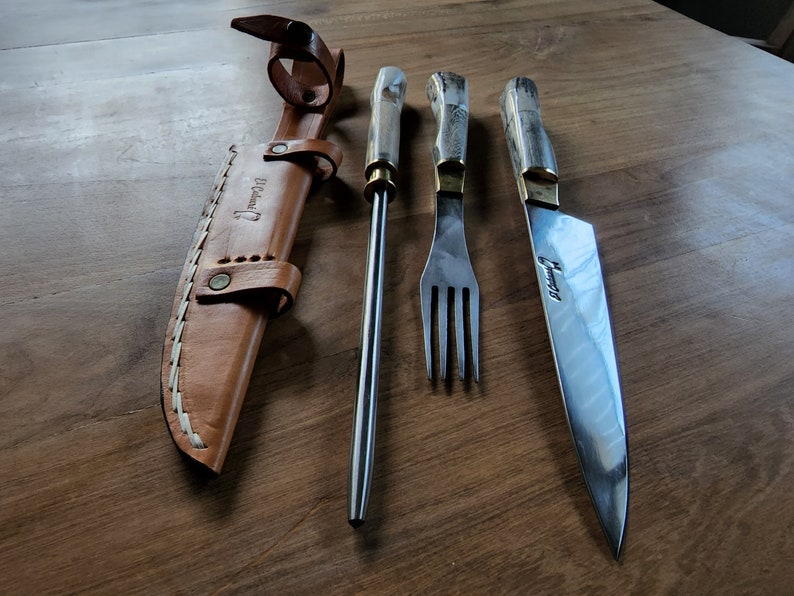 Puede incluir: Un juego de tres utensilios de cocina plateados con mangos de madera. El juego incluye un cuchillo, un tenedor y una brocheta. Los utensilios est&aacute;n dispuestos sobre una superficie de madera. El cuchillo y el tenedor tienen la palabra "Uruguay" grabada en la hoja.