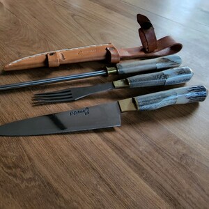 Puede incluir: Un juego de tres cuchillos con mangos de asta de ciervo y una funda de cuero. Los cuchillos incluyen un cuchillo para trinchar, un tenedor y un acero para afilar. El cuchillo para trinchar tiene el texto "St. Cuthbert" en la hoja.