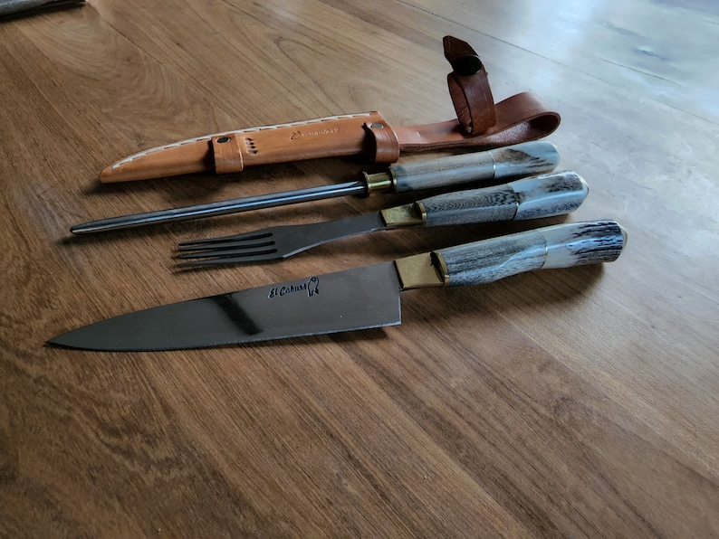Puede incluir: Un juego de tres cuchillos con mangos de asta de ciervo y una funda de cuero. Los cuchillos incluyen un cuchillo para trinchar, un tenedor y un acero para afilar. El cuchillo para trinchar tiene el texto "St. Cuthbert" en la hoja.