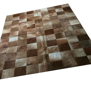 Puede incluir: Una alfombra de piel de vaca patchwork marrón y blanca con un patrón de cuadros. La alfombra está hecha de piezas individuales de piel de vaca que se han cosido juntas.