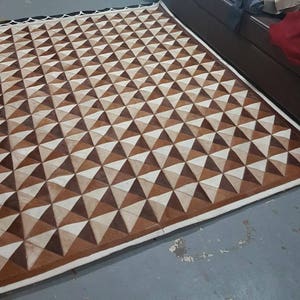May include: A brown and white geometric patterned rug made of leather. The rug has a white border and is made of many small triangles.