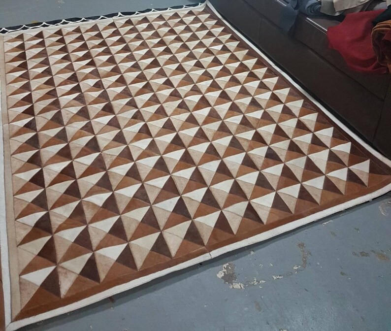 May include: A brown and white geometric patterned rug made of leather. The rug has a white border and is made of many small triangles.