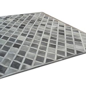 Puede incluir: Una alfombra de piel de vaca gris y blanca con un patrón de diamantes y un borde blanco. La alfombra está hecha de cuero y tiene un diseño geométrico único.