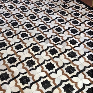 May include: A black, white and brown geometric patterned rug with a black border. The rug is made of leather and has a unique design.