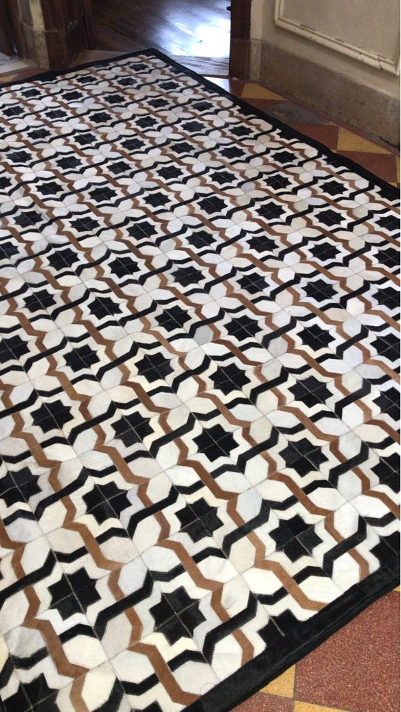 May include: A black, white and brown geometric patterned rug with a black border. The rug is made of leather and has a unique design.