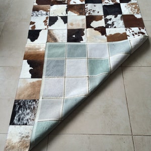 Op de afbeelding: Een patchwork vloerkleed met vierkanten van koeienhuid in bruin, zwart en wit. De onderkant van het vloerkleed is zichtbaar en toont een lichtblauw en grijs rasterpatroon. Het vloerkleed is rechthoekig.