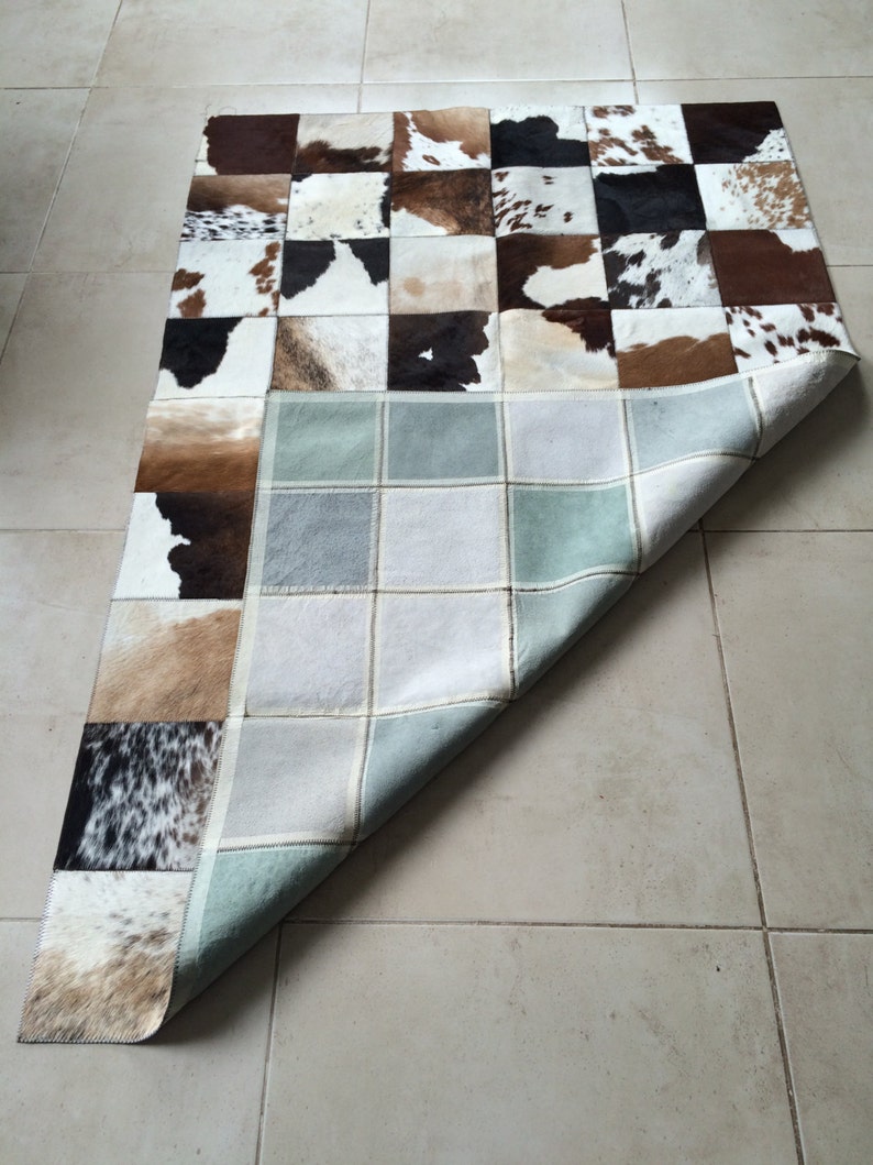 Op de afbeelding: Een patchwork vloerkleed met vierkanten van koeienhuid in bruin, zwart en wit. De onderkant van het vloerkleed is zichtbaar en toont een lichtblauw en grijs rasterpatroon. Het vloerkleed is rechthoekig.