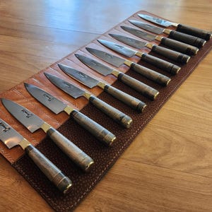 May include: A brown leather knife roll with 12 slots holds a set of sharp, silver kitchen knives. Each knife has a wooden handle and is labeled with the word "Knife" in a black font.