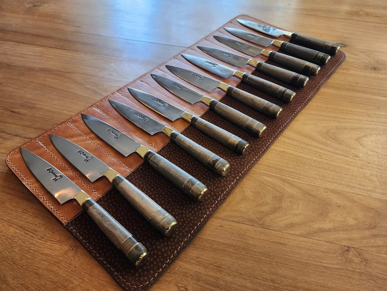 May include: A brown leather knife roll with 12 slots holds a set of sharp, silver kitchen knives. Each knife has a wooden handle and is labeled with the word "Knife" in a black font.