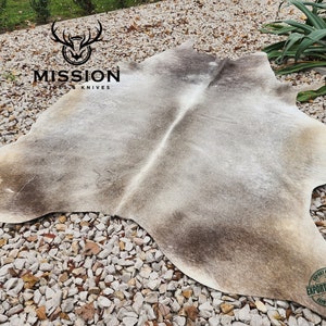May include: A large, light gray cowhide rug with a dark brown patch in the center. The rug is laid out on a bed of small, white rocks. The text "MISSION HIDES & KNIVES" is visible in the top left corner of the image. A round, red stamp with the text "EXPORT QUALITY" is visible in the bottom right corner of the image.