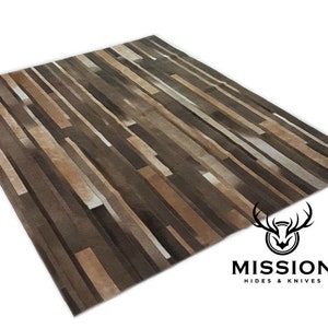Puede incluir: Una alfombra de piel de vaca marrón, beige y blanca con un patrón a rayas. La alfombra está hecha de tiras de cuero cosidas juntas. La alfombra está hecha por Mission Hides & Knives.
