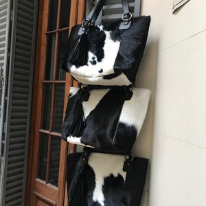 May include: Three black and white cowhide tote bags with black tassels hanging from a hook. The bags are stacked on top of each other.