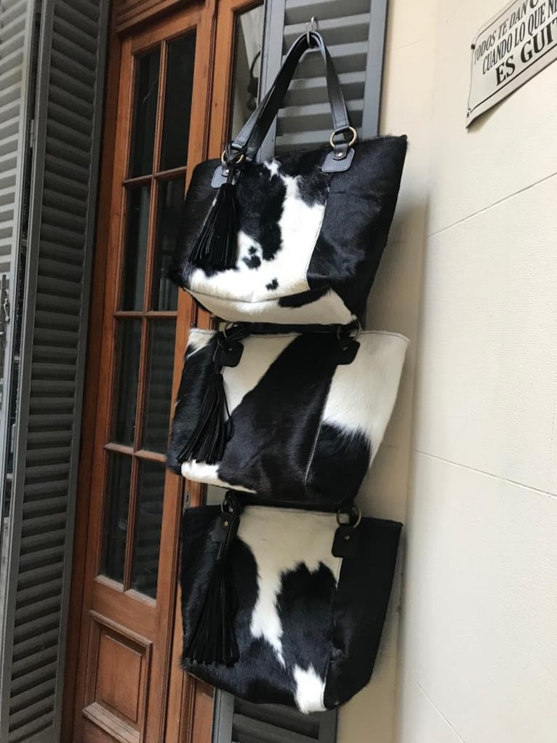 May include: Three black and white cowhide tote bags with black tassels hanging from a hook. The bags are stacked on top of each other.