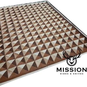 May include: A brown and white leather rug with a geometric pattern of triangles. The rug is made from cowhide and has a white border. The rug is labelled "Mission Hides & Knives".