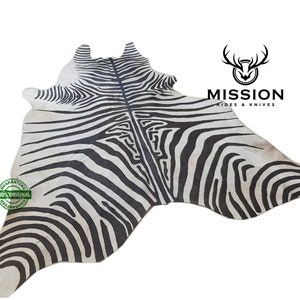 May include: Black and white zebra hide rug with a natural, slightly textured surface. The rug is large and features a distinctive zebra pattern. The rug is labeled "Mission Hides & Knives" in the upper right corner.