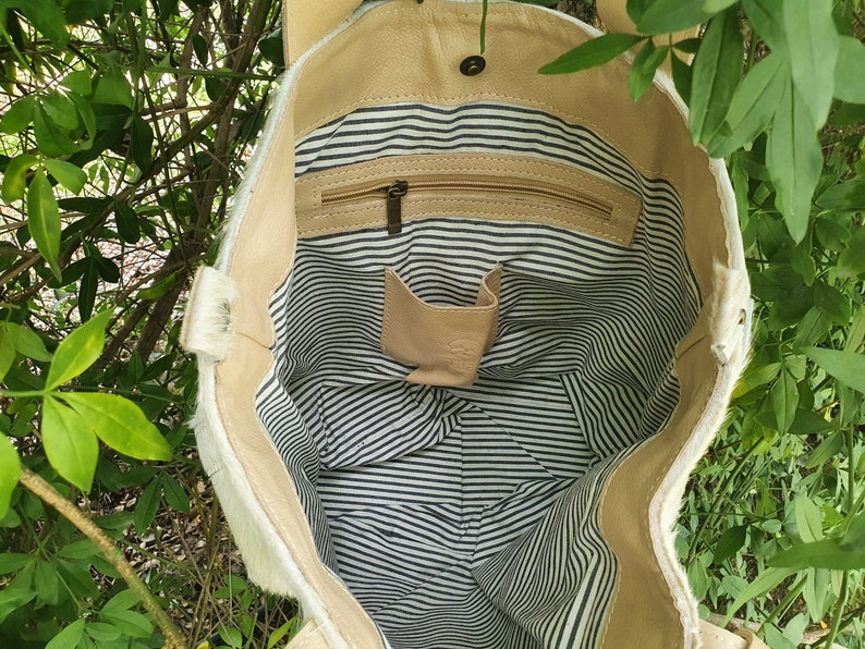 May include: A white cowhide tote bag with a tan leather lining. The bag has a zippered pocket and a small interior pocket. The lining is striped in black and white.