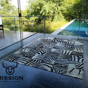 Puede incluir: Una alfombra de piel de cebra en blanco y negro hecha de piezas de cuero de patchwork. La alfombra está colocada sobre un suelo de baldosas frente a una puerta de cristal que conduce a un patio con una piscina.