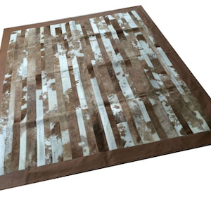May include: A brown and white cowhide rug with a brown leather border. The rug is made of strips of cowhide sewn together to create a striped pattern.