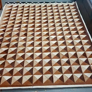 May include: A brown and white geometric patterned rug made of leather. The rug has a white border and is made up of a repeating pattern of triangles.