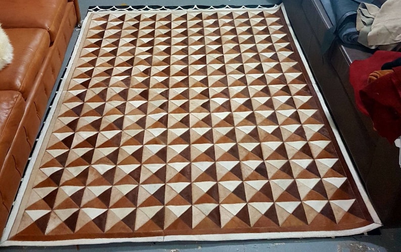 May include: A brown and white geometric patterned rug made of leather. The rug has a white border and is made up of a repeating pattern of triangles.