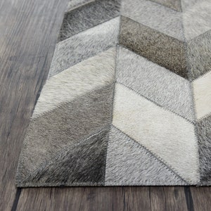 Puede incluir: Una alfombra de piel de vaca con estampado geométrico gris y blanco con un diseño cosido. La alfombra está hecha de piel de vaca natural y tiene una sensación suave y lujosa.