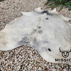 May include: A white cowhide rug with black markings. The rug is laid out on a gravel surface. The Mission Hides & Knives logo is visible in the bottom right corner.