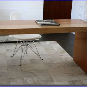 May include: A light wood desk with a white chair and a book on top. The desk has a single leg on the right side and a white rug underneath.