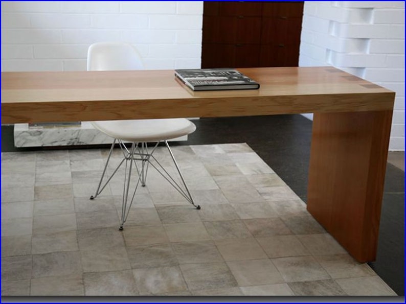 May include: A light wood desk with a white chair and a book on top. The desk has a single leg on the right side and a white rug underneath.