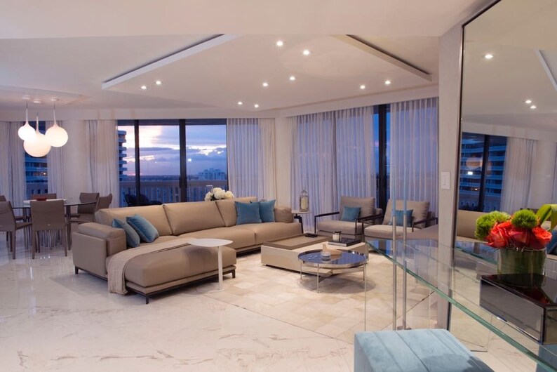 May include: A modern living room featuring a large sectional sofa, a glass coffee table, and a view of the city skyline. The room is decorated in a neutral colour palette with pops of blue.