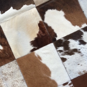 May include: A patchwork rug made from cowhide squares in various shades of brown, white, and black. The squares are stitched together, creating a textured surface. The rug's design offers a rustic aesthetic, with each square displaying unique patterns.