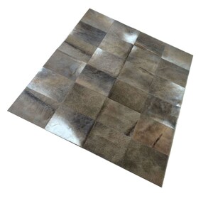 Puede incluir: Una alfombra de patchwork hecha de piel de vaca con una variedad de tonos marrones y blancos. La alfombra está hecha de cuadrados de diferentes tamaños y está dispuesta en un patrón aleatorio.