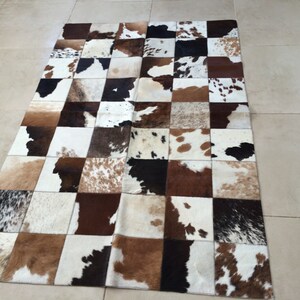 Op de afbeelding: Een rechthoekig koeienvachtkleed met een patchwork-ontwerp. Het kleed heeft vierkanten van bruin, wit en zwart koeienvel. Het kleed ligt op een lichtgekleurde tegelvloer.