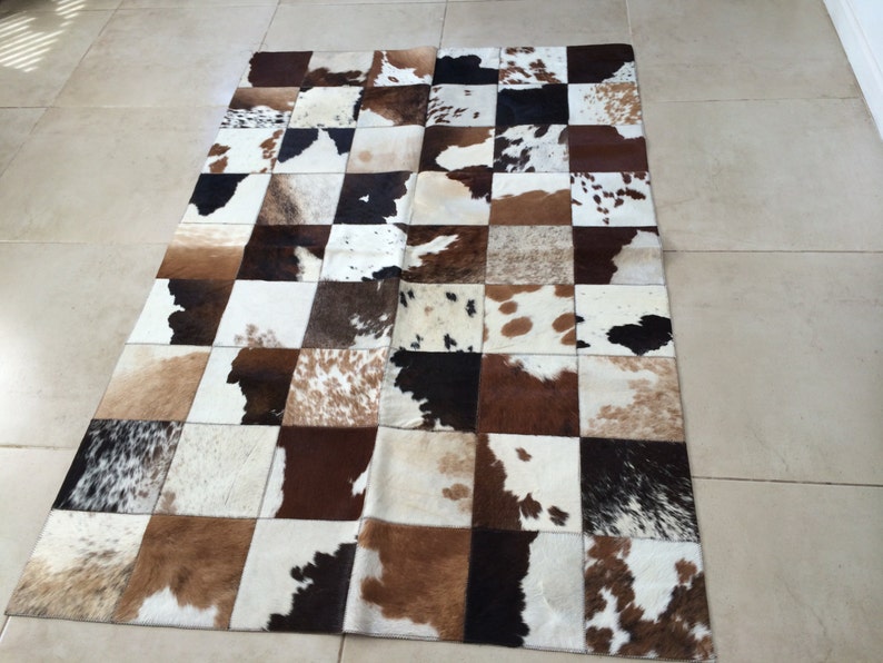 Op de afbeelding: Een rechthoekig koeienvachtkleed met een patchwork-ontwerp. Het kleed heeft vierkanten van bruin, wit en zwart koeienvel. Het kleed ligt op een lichtgekleurde tegelvloer.