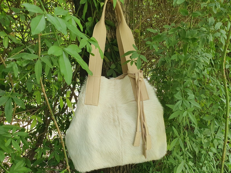 May include: A white cowhide tote bag with tan leather straps and a tassel. The bag is hanging from a tree branch.