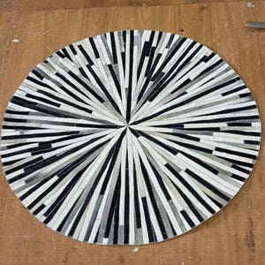 May include: A round, black and white leather rug with a geometric pattern. The rug is made of strips of leather that are arranged in a radial pattern, creating a sunburst effect.