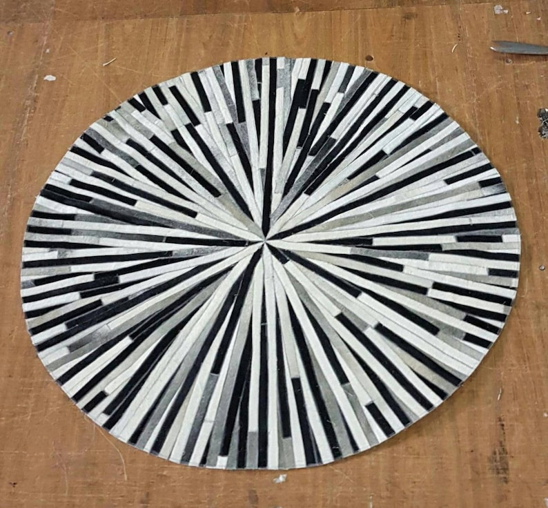 May include: A round, black and white leather rug with a geometric pattern. The rug is made of strips of leather that are arranged in a radial pattern, creating a sunburst effect.