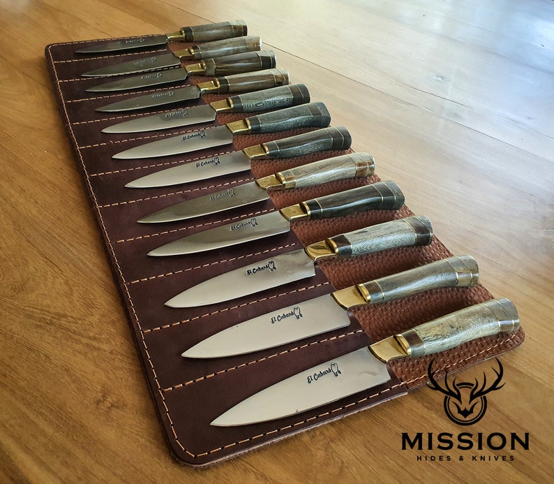 May include: A set of 12 knives with brown leather handles and brass accents are displayed in a brown leather roll with the text "MISSION HIDES & KNIVES" on the bottom right corner.