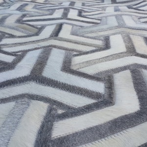 Puede incluir: Una alfombra geométrica blanca y gris hecha de piel de vaca. La alfombra tiene un diseño moderno con un patrón repetitivo de triángulos y cuadrados.