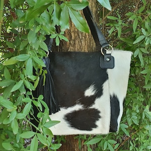 May include: A black and white cowhide tote bag with a black leather tassel and a black leather strap. The bag has a silver ring on the strap.