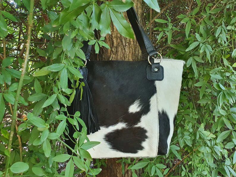 May include: A black and white cowhide tote bag with a black leather tassel and a black leather strap. The bag has a silver ring on the strap.