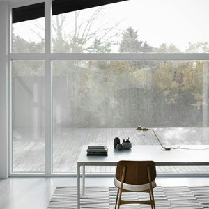 May include: A white desk with a wooden chair sits on a grey and white striped rug in front of a large window with a view of trees. A lamp with a gold base and a white shade sits on the desk, along with a stack of books and a black pencil holder.