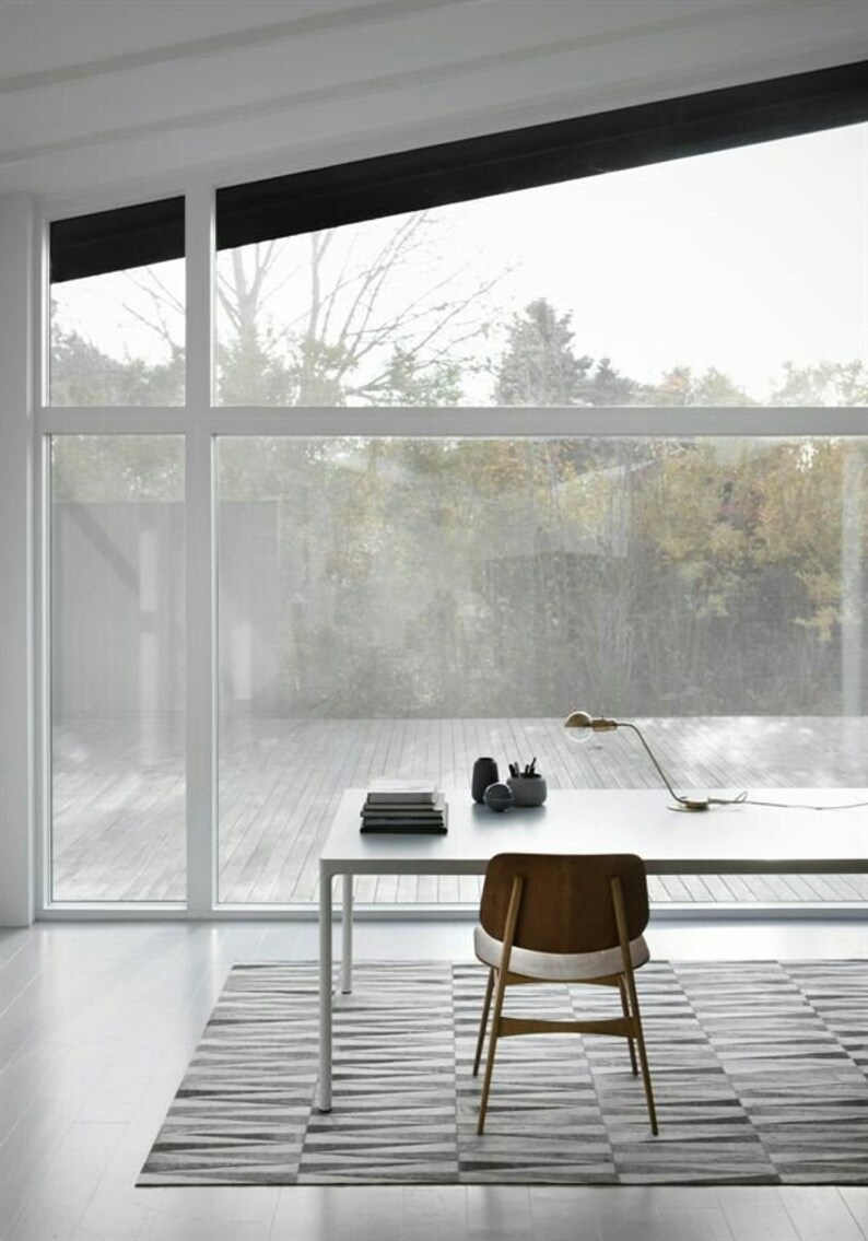 May include: A white desk with a wooden chair sits on a grey and white striped rug in front of a large window with a view of trees. A lamp with a gold base and a white shade sits on the desk, along with a stack of books and a black pencil holder.