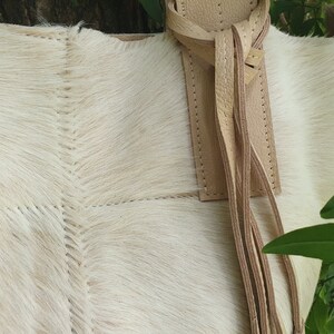 May include: A white cowhide tote bag with a tan leather strap and tassel. The bag has a stitched design and is made of soft, natural material.