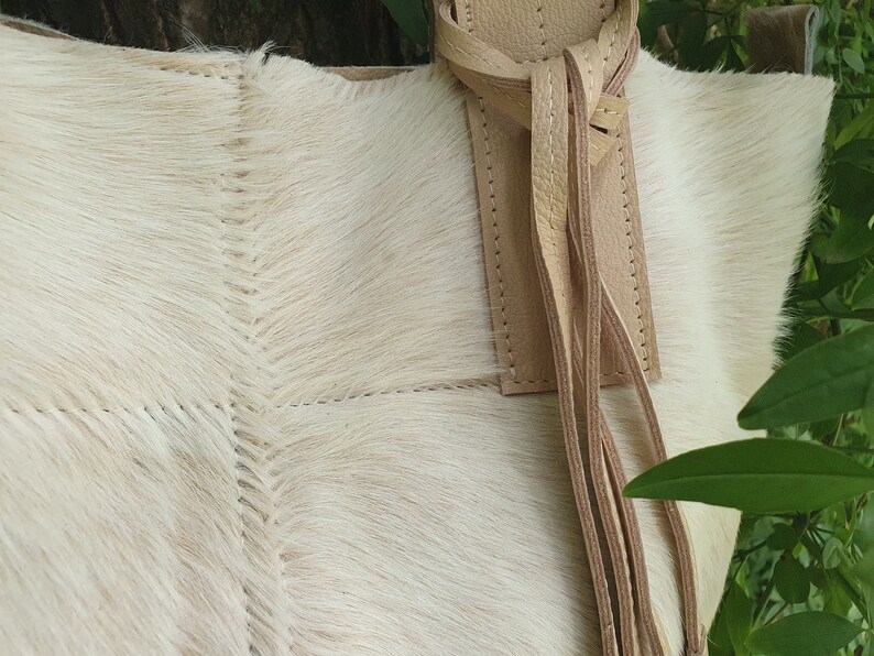 May include: A white cowhide tote bag with a tan leather strap and tassel. The bag has a stitched design and is made of soft, natural material.